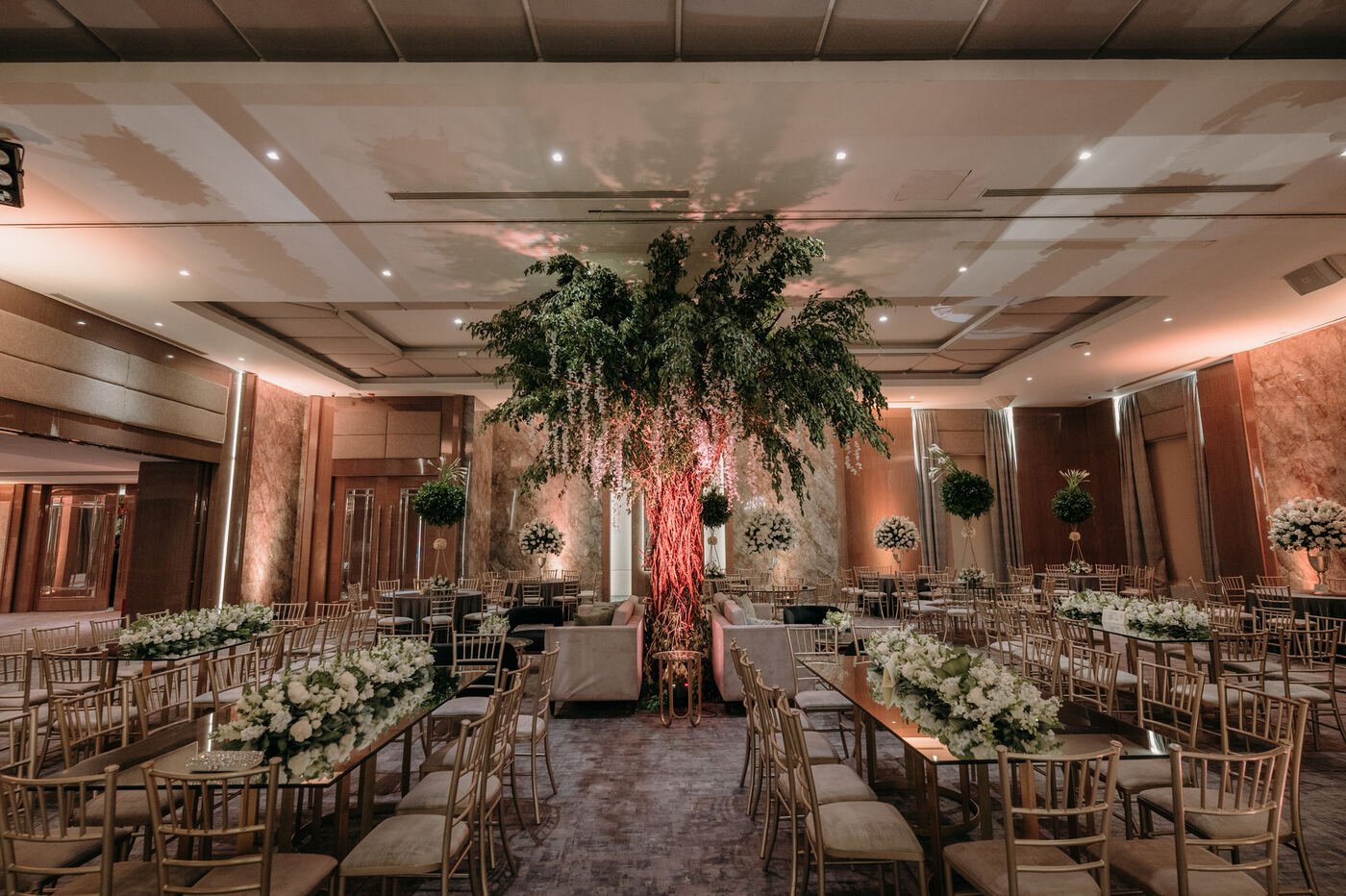 Salón montado con mesas largas, sillas doradas y centros florales
